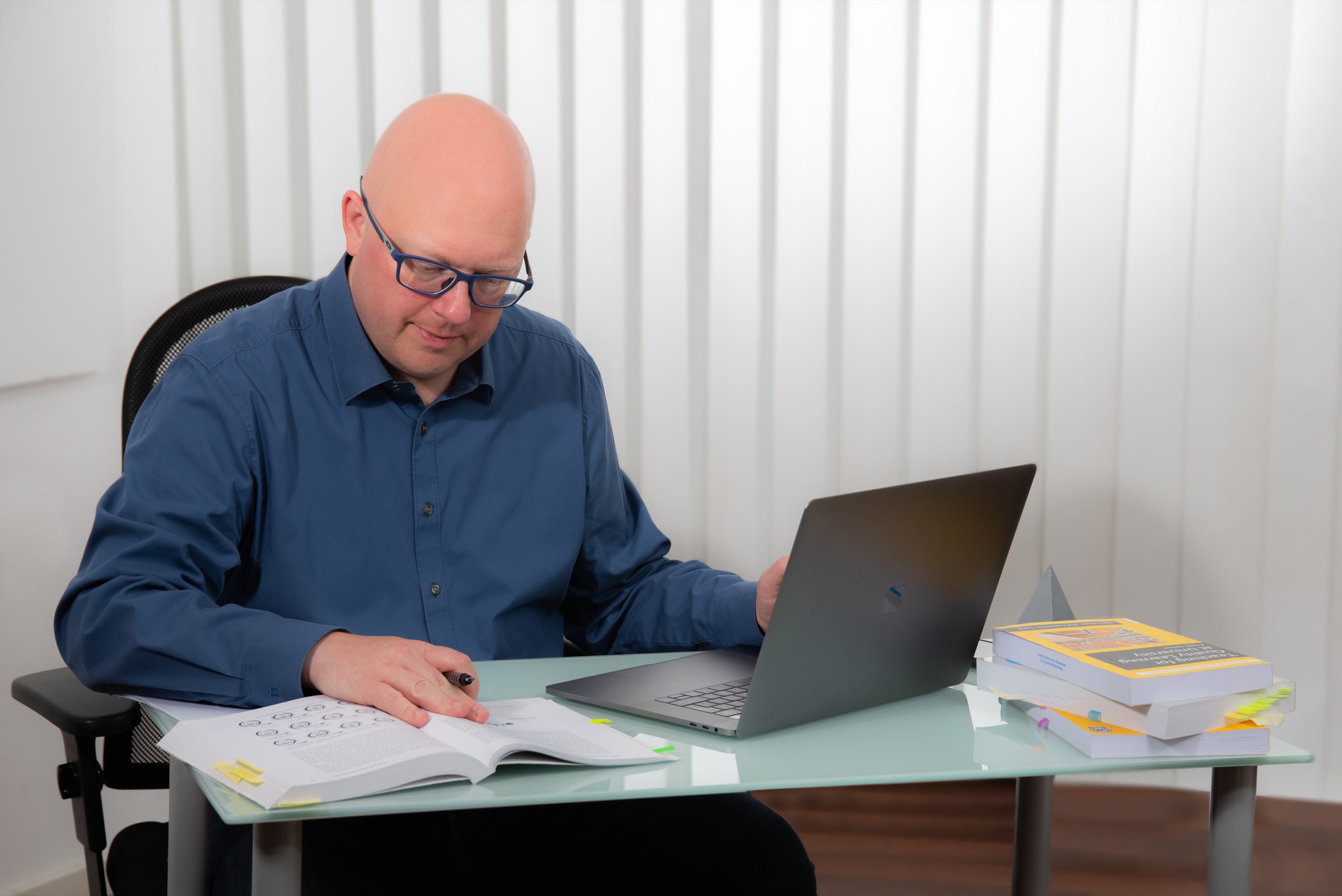 Thorsten Jungmann, ein Mann mit kurzen Haaren, dunkler Brille und dunkelblauem Hemd, sitzt am Schreibtisch vor einem Computer und lies in einem Buch.