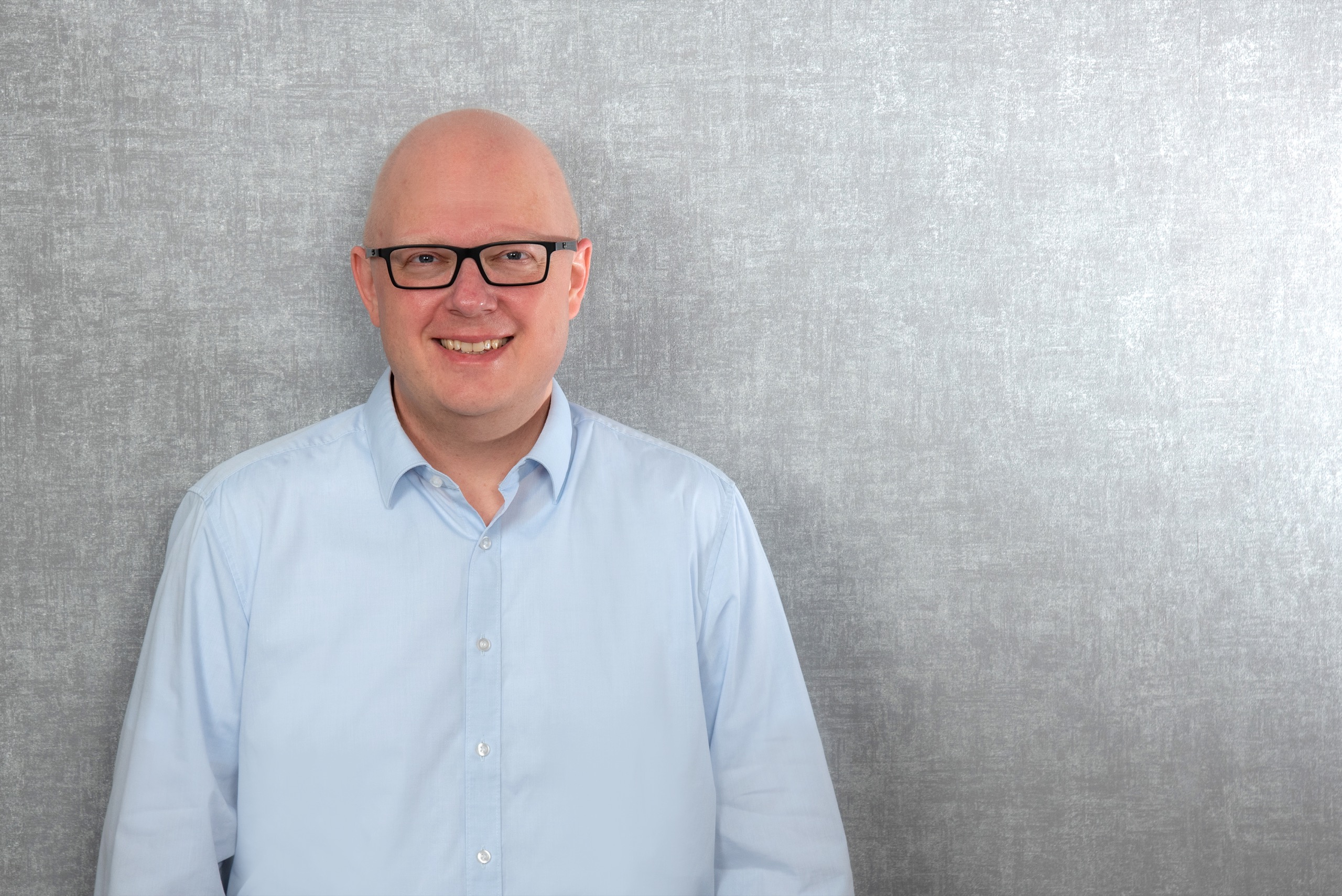 Thorsten Jungmann, ein Mann mit dunkler Brille und hellblauem Hemd vor grauem Fotohintergrund